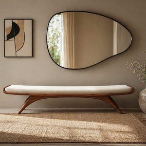 May include: An interior scene with a wooden bench, cream cushion, and an organic-shaped mirror. A framed abstract artwork hangs on the wall. A woven rug and a vase with dried flowers add to the room's aesthetic.