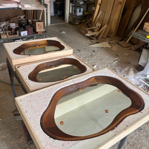 May include: Three mirrors with irregular, organic-shaped dark wood frames, each set into a light-colored wooden box filled with white packing foam. The mirrors feature smooth, curved edges and reflective surfaces, displayed in a workshop setting.