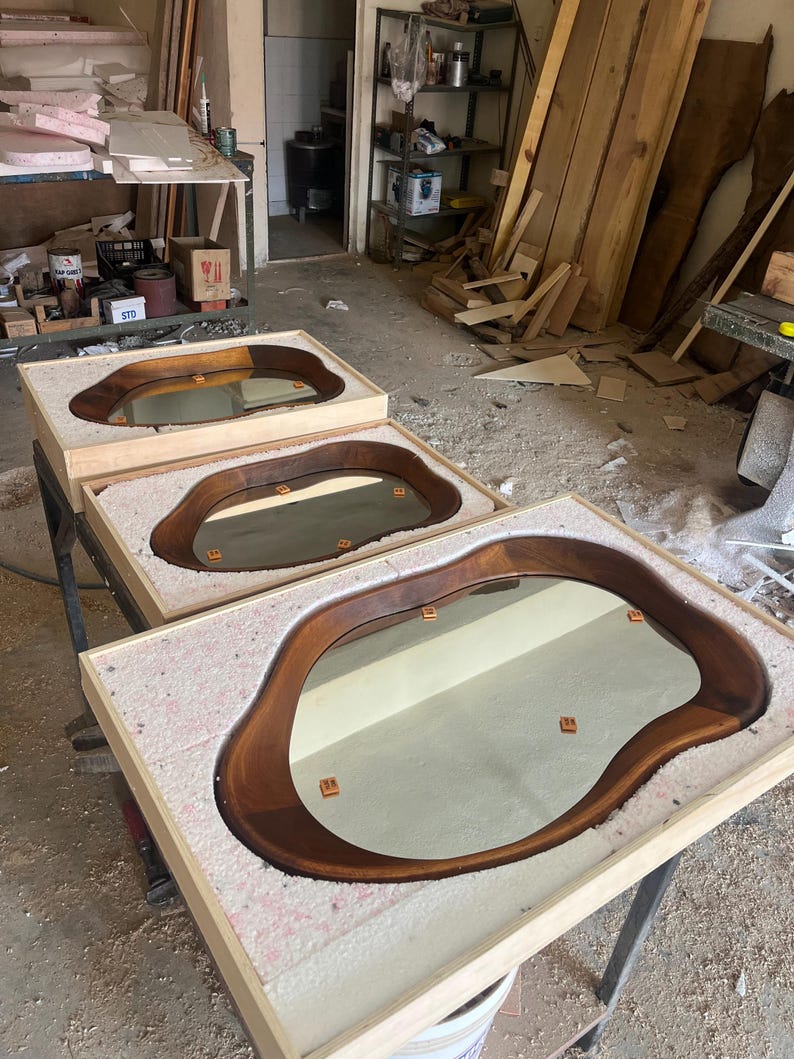 May include: Three mirrors with irregular, organic-shaped dark wood frames, each set into a light-colored wooden box filled with white packing foam. The mirrors feature smooth, curved edges and reflective surfaces, displayed in a workshop setting.