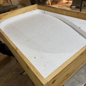 May include: A rectangular wooden frame constructed from light-colored pine boards, containing a white expanded polystyrene foam insert. The foam features a curved, raised interior border that creates a rounded shape within the rectangular structure.