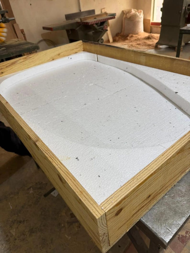 May include: A rectangular wooden frame constructed from light-colored pine boards, containing a white expanded polystyrene foam insert. The foam features a curved, raised interior border that creates a rounded shape within the rectangular structure.