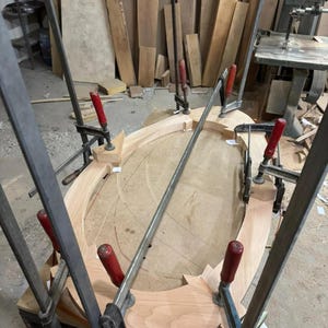 May include: An oval-shaped light-colored wooden frame is held together by several metal bar clamps with red handles. The wooden pieces are arranged in a curved structure on a flat surface, secured tightly by the clamps during the assembly process.