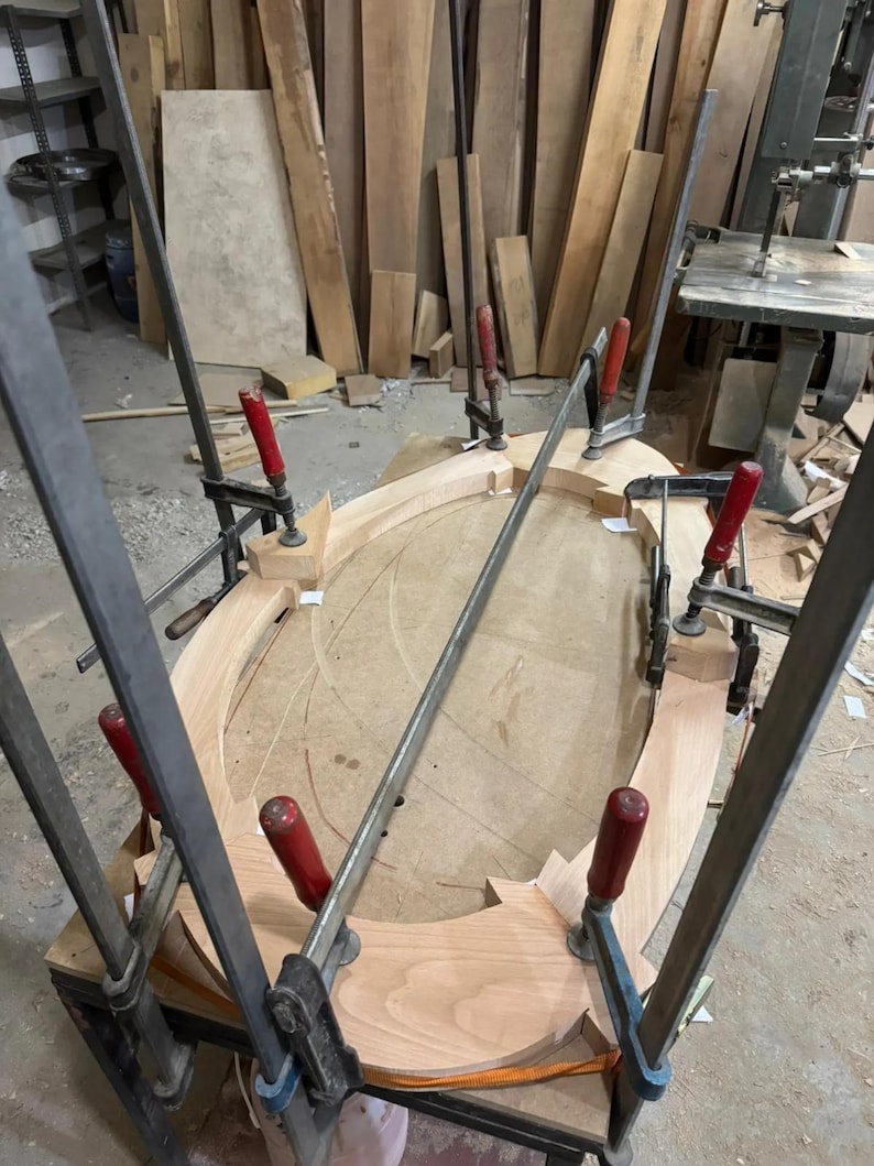 May include: An oval-shaped light-colored wooden frame is held together by several metal bar clamps with red handles. The wooden pieces are arranged in a curved structure on a flat surface, secured tightly by the clamps during the assembly process.