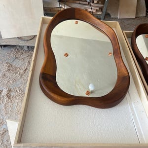 May include: A wall mirror with an irregular, organic-shaped wooden frame featuring a rich, warm brown wood grain finish. The reflective glass surface is smooth and clear, showing small square markers placed on the mirror face for alignment.