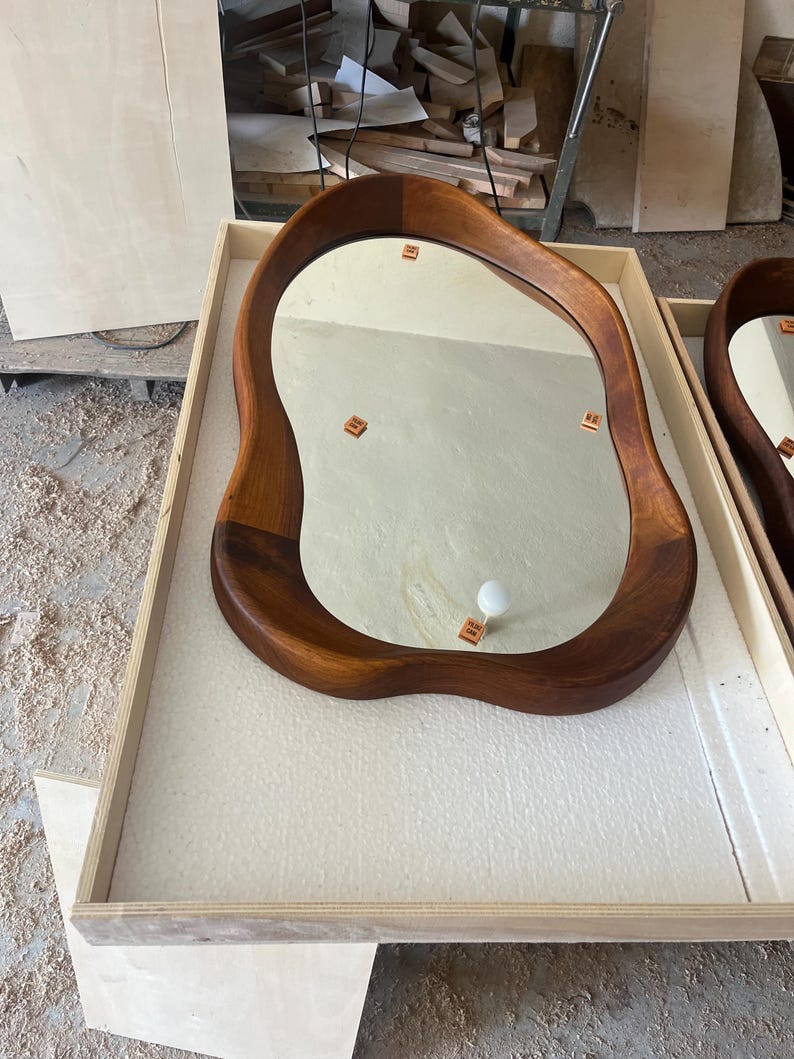 May include: A wall mirror with an irregular, organic-shaped wooden frame featuring a rich, warm brown wood grain finish. The reflective glass surface is smooth and clear, showing small square markers placed on the mirror face for alignment.