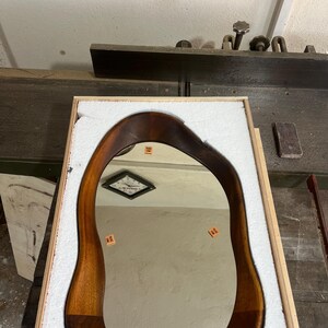 May include: An irregular, organic-shaped mirror with a dark brown wooden frame. The reflective surface features four small orange rectangular stickers with the text YILDIZ CAM printed on them. The mirror is nestled inside a protective wooden crate.