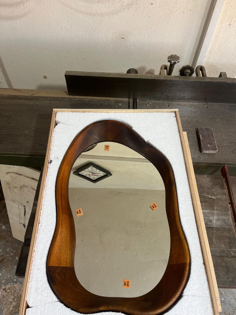 May include: An irregular, organic-shaped mirror with a dark brown wooden frame. The reflective surface features four small orange rectangular stickers with the text YILDIZ CAM printed on them. The mirror is nestled inside a protective wooden crate.