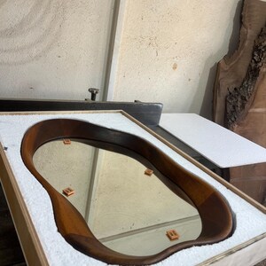 May include: An oval-shaped mirror with a smooth, dark brown wooden frame featuring an irregular, organic edge. The mirror is securely nestled within a rectangular wooden shipping crate lined with white protective foam padding for safe transport.
