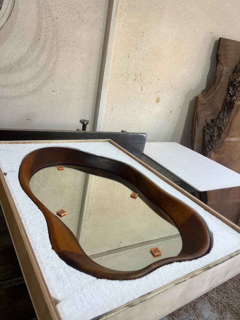 May include: An oval-shaped mirror with a smooth, dark brown wooden frame featuring an irregular, organic edge. The mirror is securely nestled within a rectangular wooden shipping crate lined with white protective foam padding for safe transport.