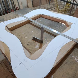 May include: A wooden frame structure featuring a white paper template with a wavy, organic outline placed on top. A long metal ruler spans across the frame diagonally, and a small metal square tool sits upright in the center of the wooden frame.