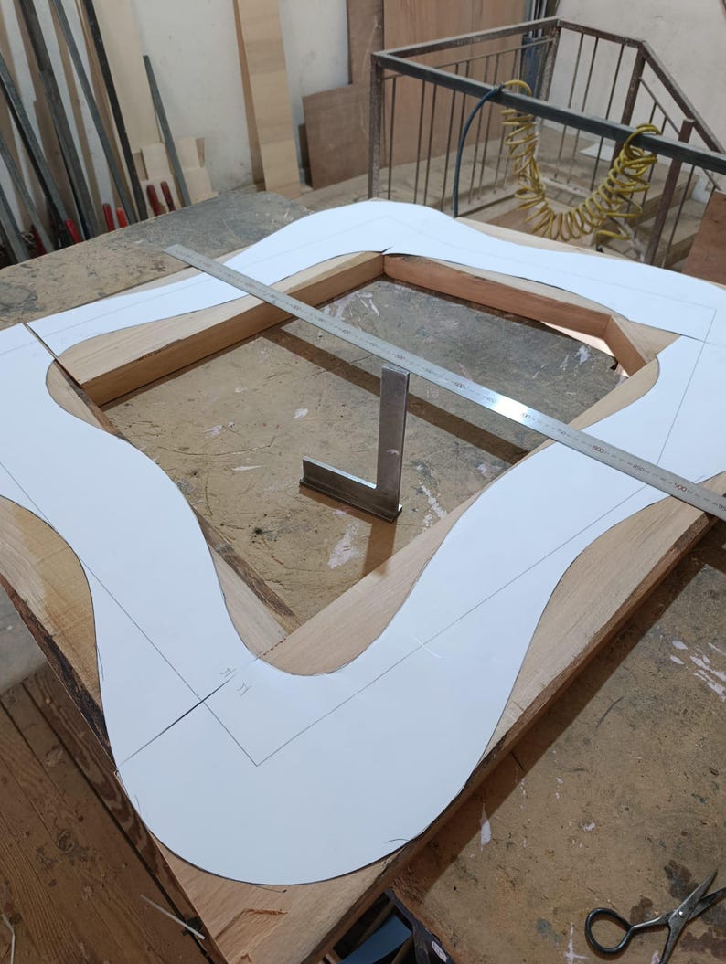 May include: A wooden frame structure featuring a white paper template with a wavy, organic outline placed on top. A long metal ruler spans across the frame diagonally, and a small metal square tool sits upright in the center of the wooden frame.