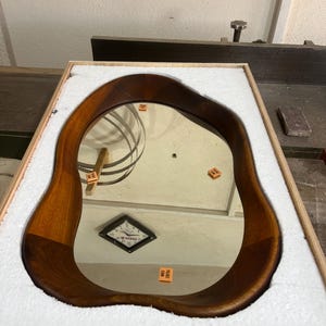 May include: An irregular, organic-shaped wall mirror featuring a smooth, dark-toned wooden frame. The mirror is securely nestled within a rectangular wooden crate lined with white protective foam padding, reflecting a clock and small orange labels.