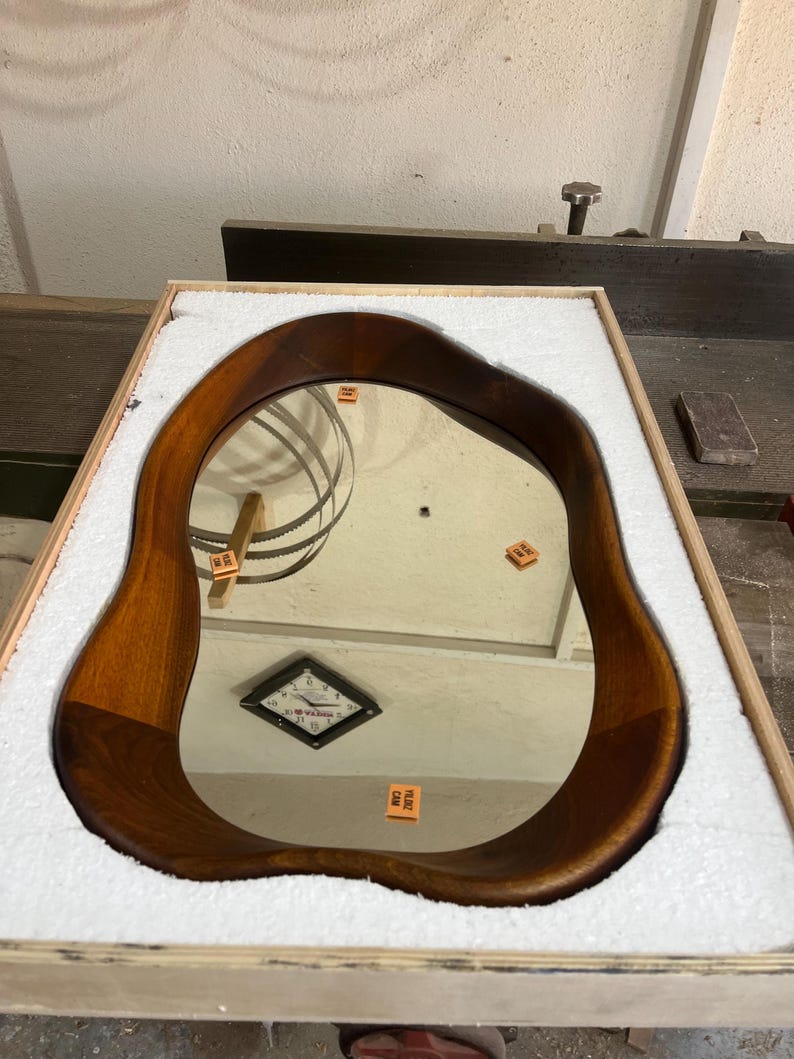 May include: An irregular, organic-shaped wall mirror featuring a smooth, dark-toned wooden frame. The mirror is securely nestled within a rectangular wooden crate lined with white protective foam padding, reflecting a clock and small orange labels.