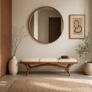 May include: A wooden bench with a cream-colored upholstered seat, positioned below a large circular mirror with a wooden frame. The scene includes a tall beige vase with branches, a framed abstract line drawing, and a small bowl on the bench.