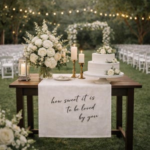 May include: A wooden table displays a two-tiered white wedding cake, floral arrangements, and lit candles. A white runner with the text "how sweet it is to be loved by you" is draped across the table. The scene is set outdoors with fairy lights.
