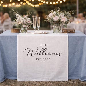 May include: A table set for a wedding reception, covered with a light blue tablecloth and a white linen runner. The runner features the text "The Williams EST. 2025" in elegant script. Floral arrangements and champagne flutes adorn the table.