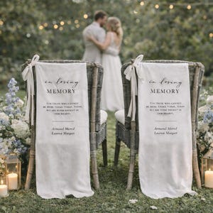 Pode incluir: Duas cadeiras de vime com tecido branco drapeado nas costas, cada uma com o texto "In Loving Memory". As cadeiras estão montadas para um casamento, com arranjos florais e velas de cada lado. Um casal está desfocado ao fundo.