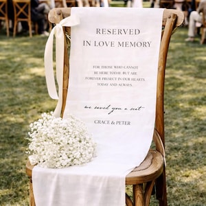 Pode incluir: Cadeira de madeira com tecido branco com a frase "RESERVED IN LOVE MEMORY" e "we saved you a seat". Um buquê de flores brancas adorna o tecido, ideal para um evento comemorativo.