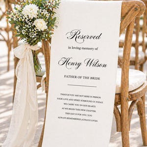 Pode incluir: Uma cadeira de madeira com uma faixa branca que diz "Reserved in loving memory of Henry Wilson, Father of the Bride". Um buquê de flores brancas e vegetação está amarrado à cadeira.