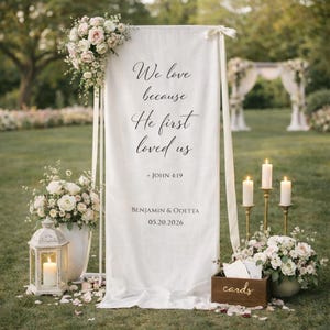 May include: A white wedding sign with the text "We love because He first loved us" and the names Benjamin & Odetta, with the date 05.20.2026. The sign is decorated with flowers and candles, set outdoors.