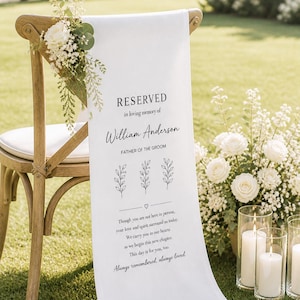 Pode incluir: Cadeira de madeira com tecido branco drapeado no encosto, adornada com arranjos florais. O tecido exibe o texto "RESERVED" e uma mensagem memorial para William Anderson, Pai do Noivo, com elementos florais.