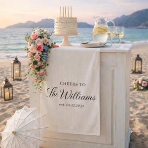 Puede incluir: Una recepción de boda en la playa con pastel, flores y bebidas. Un pastel blanco con velas está sobre un soporte rosa. Una pancarta dice "Cheers to The Williams" con la fecha 08.12.2025.