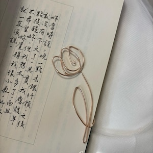 Peut inclure: Un marque-page en fil métallique couleur or rose en forme de tulipe avec une seule feuille, posé sur une page de livre ouverte avec du texte chinois vertical noir et les mots Karina The Precious imprimés en bas de page.