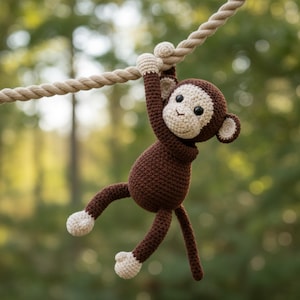 May include: A handmade crochet monkey in shades of brown and cream, hanging from a thick rope. The monkey has a light brown face, ears, and hands, with dark brown body and limbs. The background is a blurred view of green trees.