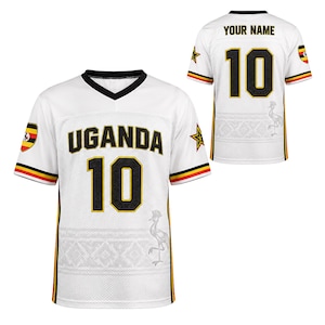 May include: A white V-neck jersey featuring the word UGANDA and the number 10 in black with gold outlines. The sleeves have black, yellow, and red stripes, and the fabric displays a subtle grey crane graphic and geometric patterns on the lower section.