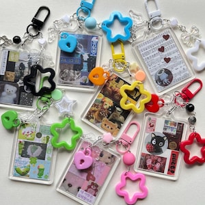 May include: A selection of clear acrylic keyrings with diverse charms and images. The keyrings feature star, heart, and flower shapes in colours like pink, green, yellow, and blue. Some keyrings have images of bears, food, and text.