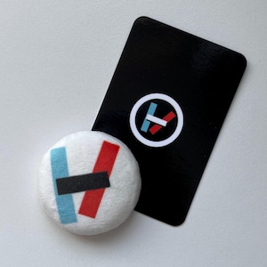 Puede incluir: Un objeto redondo de felpa blanca con un gráfico de una barra vertical azul claro, una barra vertical roja y una barra horizontal negra. Junto a él hay una tarjeta rectangular negra con un logotipo circular blanco que contiene barras de colores.