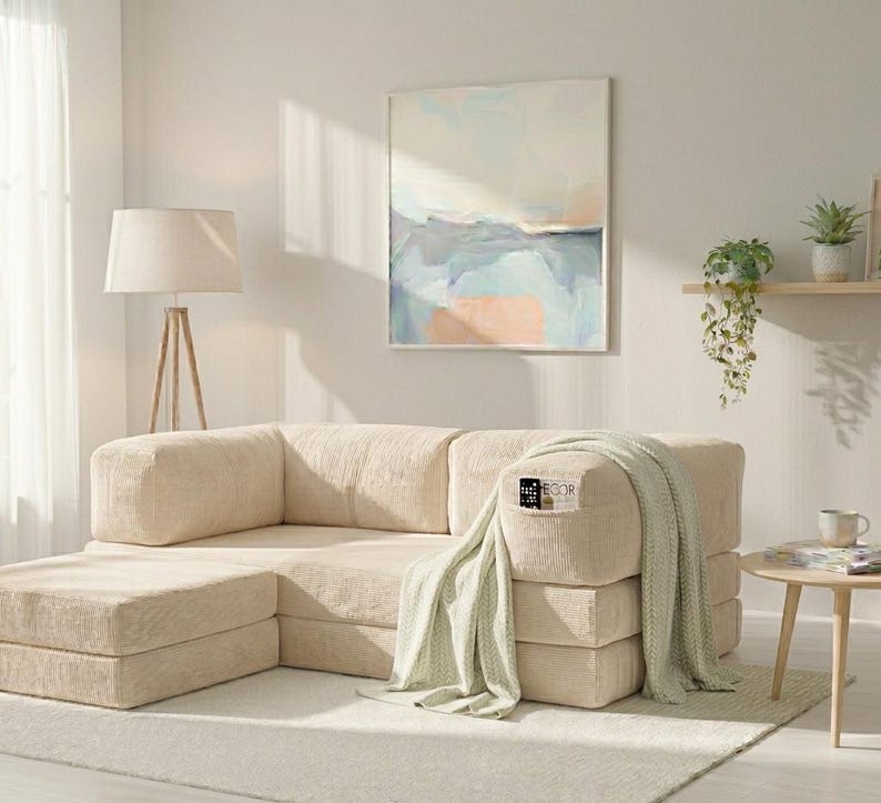 May include: A modular beige corduroy sofa with a matching ottoman, featuring a textured fabric finish. A light green knit throw blanket is draped over one armrest, and a small black remote control is tucked into the side pocket of the sofa.