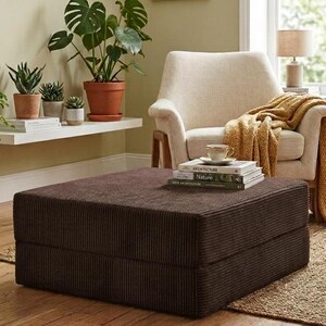 Pu&ograve; includere: Un pouf quadrato in velluto a coste marrone scuro con una trama rigata. Il pouf &egrave; posizionato davanti a una poltrona imbottita color bianco sporco, drappeggiata con una coperta in maglia color senape e sormontata da libri e una tazza.