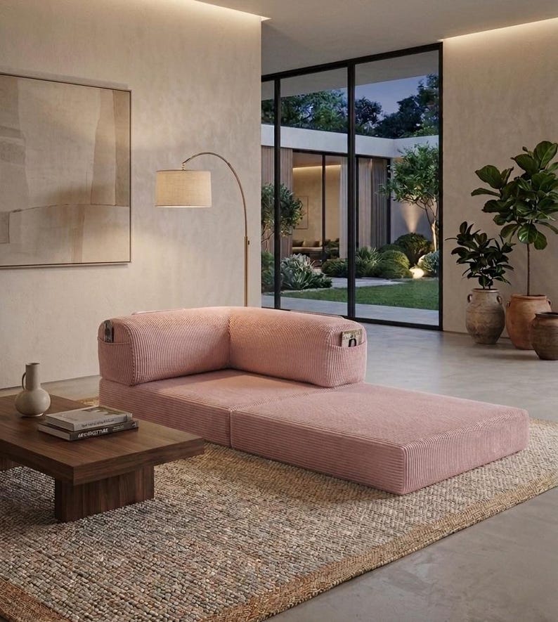 May include: A low-profile, pink corduroy modular sofa with a corner backrest and integrated side pockets. The sofa features a textured fabric finish and a rectangular shape, positioned on a woven rug in a room with a wooden coffee table and floor lamp.