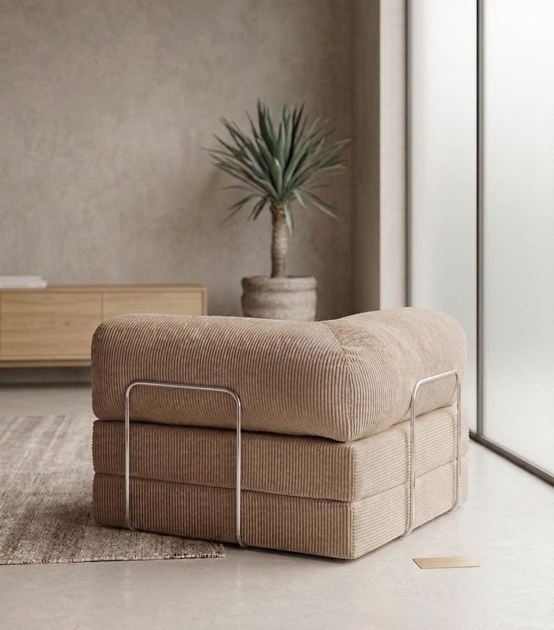 May include: A low-profile modular armchair upholstered in beige corduroy fabric. The seat features a thick, square cushion design supported by a sleek, silver-toned metal frame that wraps around the sides and back of the furniture piece.