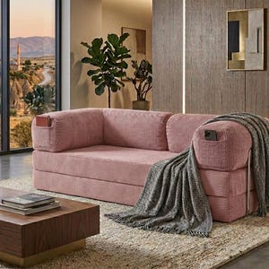 May include: A pink corduroy sofa featuring integrated side pockets on the armrests, one containing a small book and the other a smartphone. A grey textured throw blanket is draped over the right armrest of the sofa, which has a low-profile design.