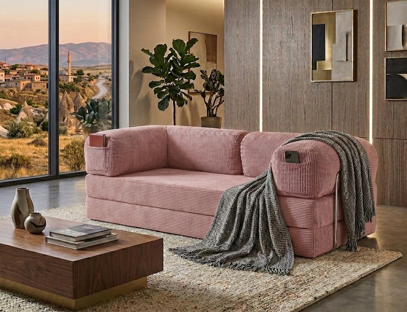 May include: A pink corduroy sofa featuring integrated side pockets on the armrests, one containing a small book and the other a smartphone. A grey textured throw blanket is draped over the right armrest of the sofa, which has a low-profile design.