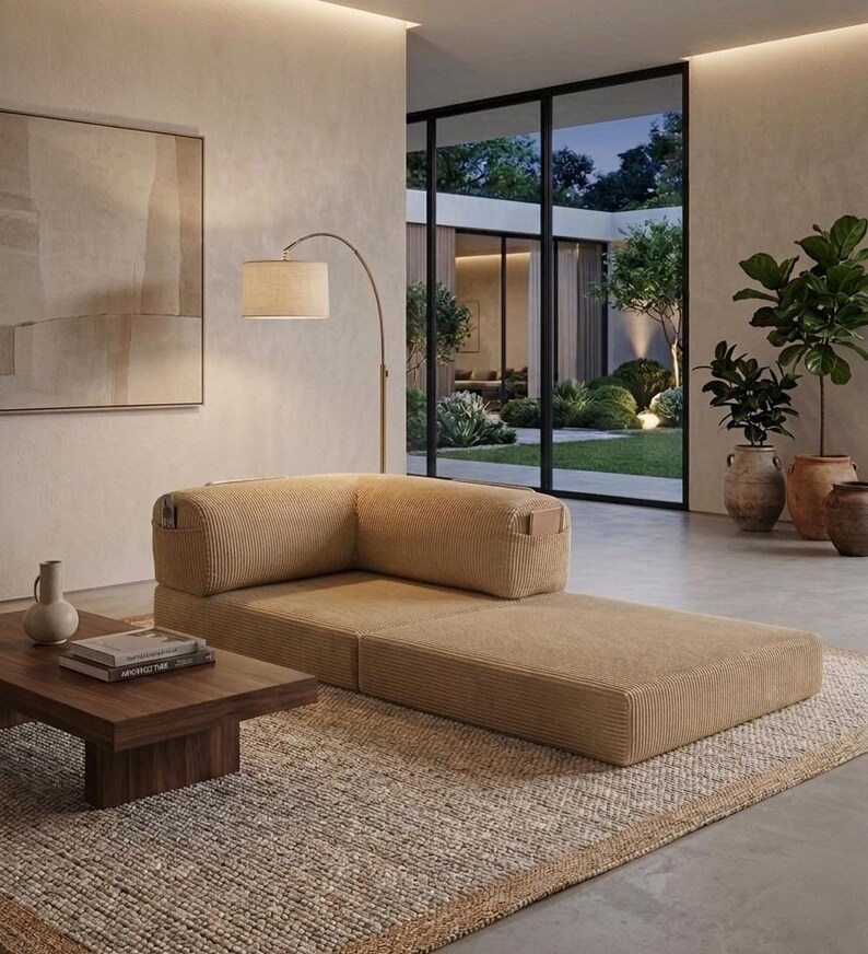 May include: A low-profile, tan corduroy modular sofa with a corner backrest and integrated side pockets, positioned on a textured rug next to a dark wood coffee table holding a small vase and stacked books, illuminated by a curved floor lamp.