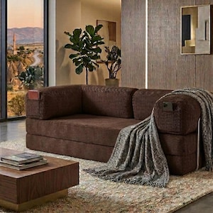 Pu&ograve; includere: Un divano in velluto a coste marrone scuro dal design a profilo basso con tasche laterali integrate sui braccioli. Una coperta testurizzata grigia e bianca &egrave; drappeggiata sul lato destro del divano, posizionato su un tappeto chiaro.