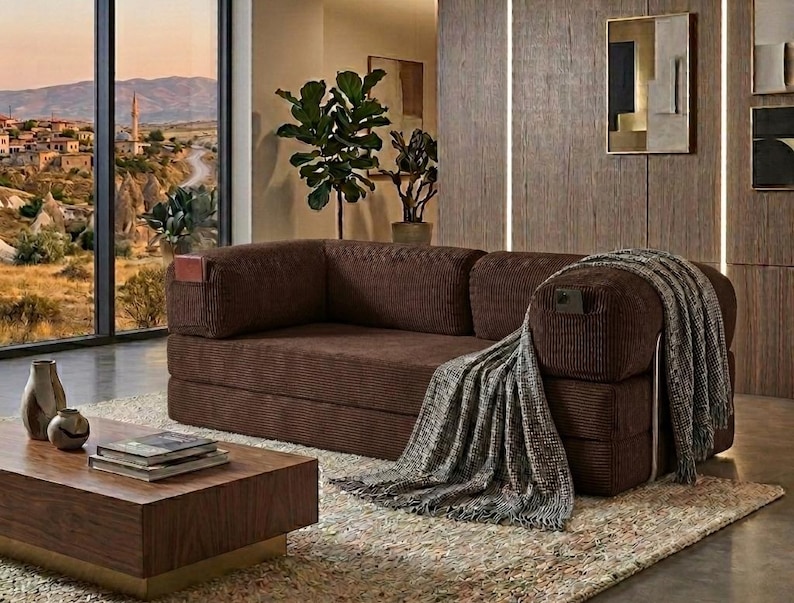 Pu&ograve; includere: Un divano in velluto a coste marrone scuro dal design a profilo basso con tasche laterali integrate sui braccioli. Una coperta testurizzata grigia e bianca &egrave; drappeggiata sul lato destro del divano, posizionato su un tappeto chiaro.