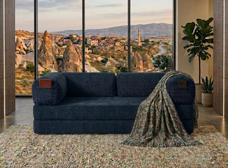 May include: A dark blue corduroy sofa with a rectangular brown leather accent on each armrest. A textured, multi-colored throw blanket with fringe edges is draped over the right side of the sofa, covering the armrest and part of the seat cushion.