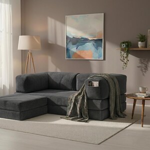 May include: A dark gray corduroy modular sofa with a matching ottoman and a built-in side pocket. A textured green throw blanket is draped over the armrest, and the sofa features a low-profile, blocky design with thick, cushioned seating sections.