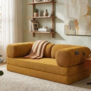 May include: A mustard yellow corduroy sofa with a low profile, rounded armrests, and a built-in side pocket containing a smartphone and keys. A beige and white checkered throw blanket is draped over the center of the seat cushions.