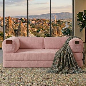 May include: A pink corduroy sofa featuring a low-profile design with two rectangular cushions and rounded armrests. Each armrest is accented with a small, dark brown rectangular leather-like patch. A dark green patterned throw blanket is draped over one side.