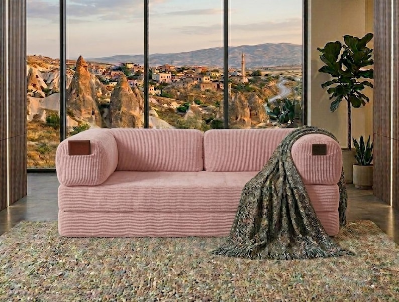 May include: A pink corduroy sofa featuring a low-profile design with two rectangular cushions and rounded armrests. Each armrest is accented with a small, dark brown rectangular leather-like patch. A dark green patterned throw blanket is draped over one side.