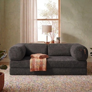 May include: A dark gray corduroy sofa featuring a low-profile design with rounded bolster-style armrests. A folded, textured orange and yellow patterned throw blanket rests on the seat cushion, adding a touch of color to the dark fabric.