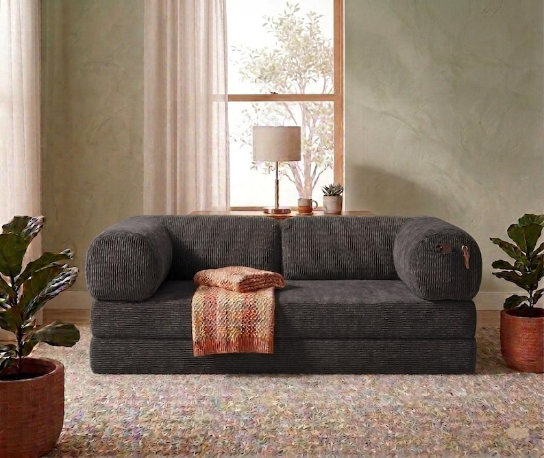 May include: A dark gray corduroy sofa featuring a low-profile design with rounded bolster-style armrests. A folded, textured orange and yellow patterned throw blanket rests on the seat cushion, adding a touch of color to the dark fabric.