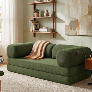 May include: A dark green corduroy sofa featuring a low-profile design with rounded armrests, a textured fabric finish, and a small side pocket on the right armrest containing a smartphone and keys, draped with a tan and white checkered throw blanket.