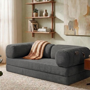 May include: A dark gray corduroy convertible sofa bed featuring a low-profile design with rounded bolster armrests. The sofa includes a side pocket for storage and is draped with a textured, light orange woven throw blanket across the seat.