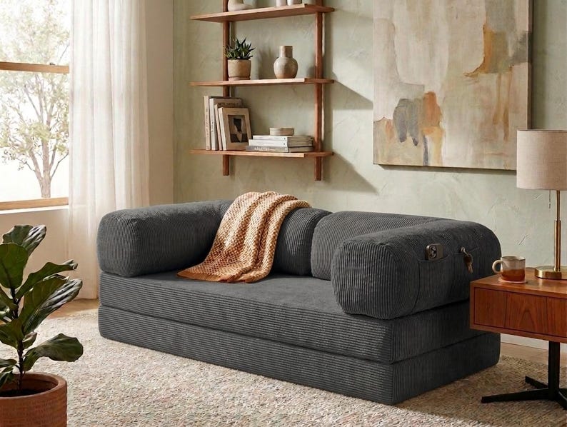 May include: A dark gray corduroy convertible sofa bed featuring a low-profile design with rounded bolster armrests. The sofa includes a side pocket for storage and is draped with a textured, light orange woven throw blanket across the seat.
